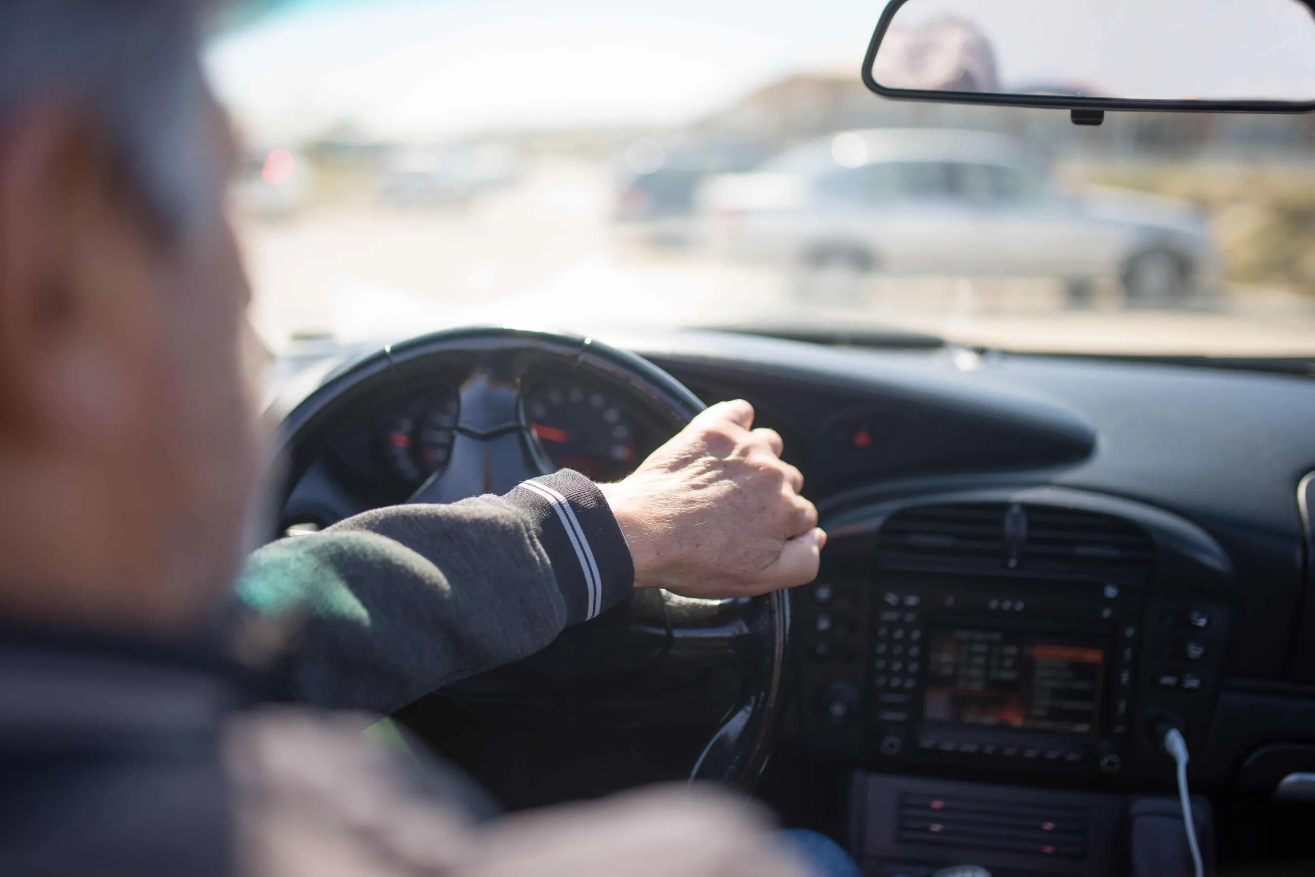 Person driving a car, viewed from the passenger seat, with one hand on the steering wheel and a blurry outdoor scene visible through the windshield. - Ghassemi Law Group Person driving a car, viewed from the passenger seat, with one hand on the steering wheel and a blurry outdoor scene visible through the windshield