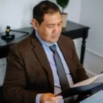 A man in a suit sits at a desk, reading documents from a folder. There is a gavel, files, and a plant on the black table behind him. - Ghassemi Law Group A man in a suit sits at a desk, reading documents from a folder There is a gavel, files, and a plant on the black table behind him