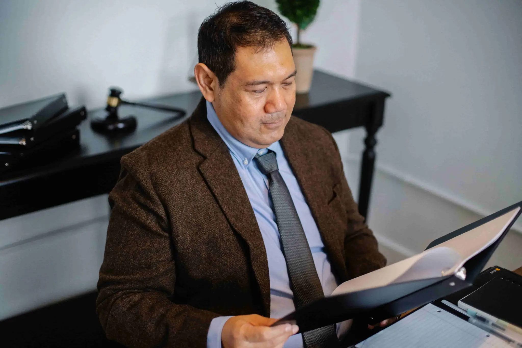 A man in a suit sits at a desk, reading documents from a folder. There is a gavel, files, and a plant on the black table behind him. - Ghassemi Law Group A man in a suit sits at a desk, reading documents from a folder There is a gavel, files, and a plant on the black table behind him