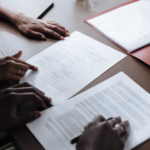 Two people sit at a desk reviewing and signing documents, with a red folder and pen nearby. - Ghassemi Law Group Two people sit at a desk reviewing and signing documents, with a red folder and pen nearby