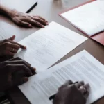 Two people are reviewing and signing documents on a desk with a red folder and pen visible nearby 