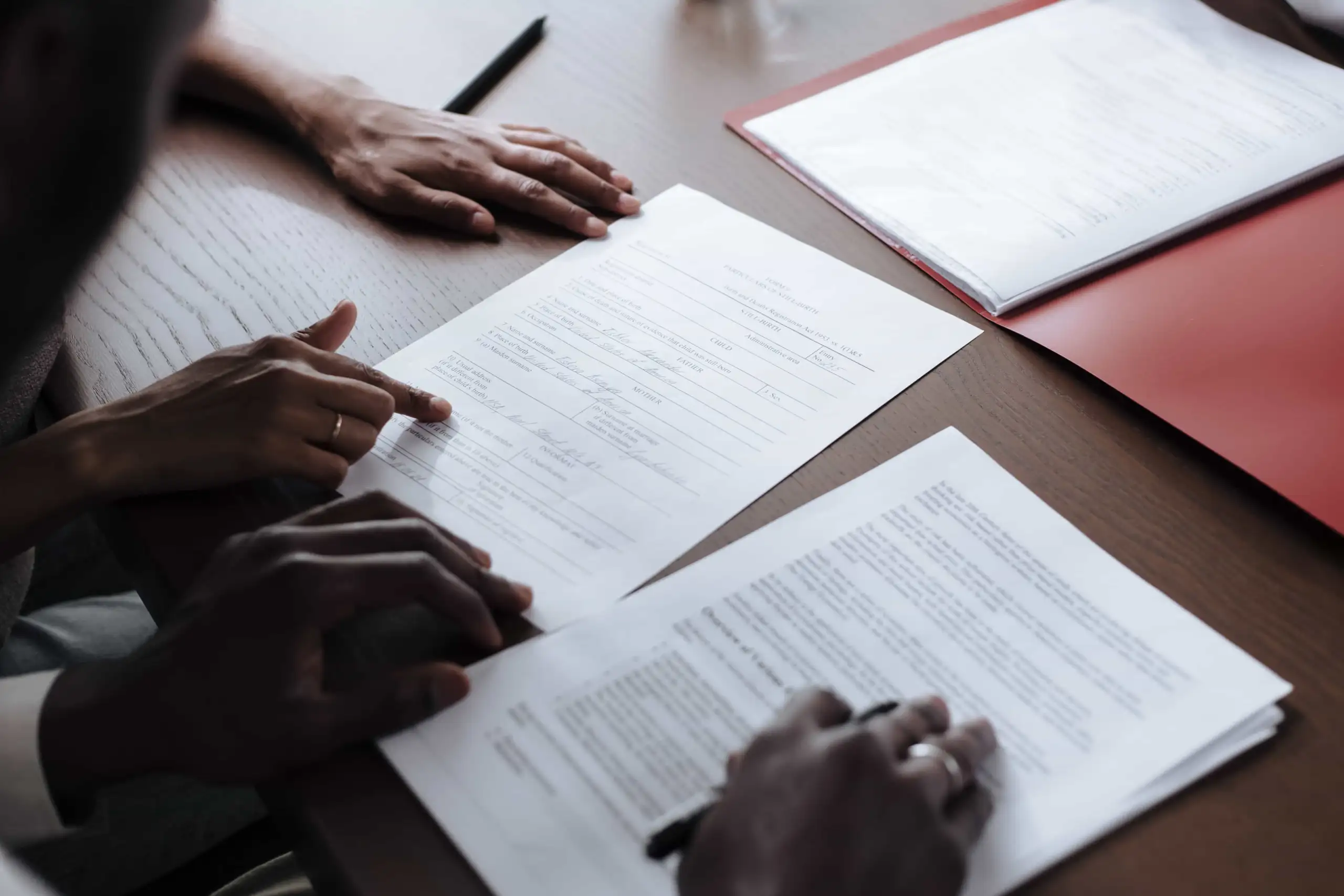 Two people are reviewing and signing documents on a desk with a red folder and pen visible nearby. - Ghassemi Law Group Two people are reviewing and signing documents on a desk with a red folder and pen visible nearby