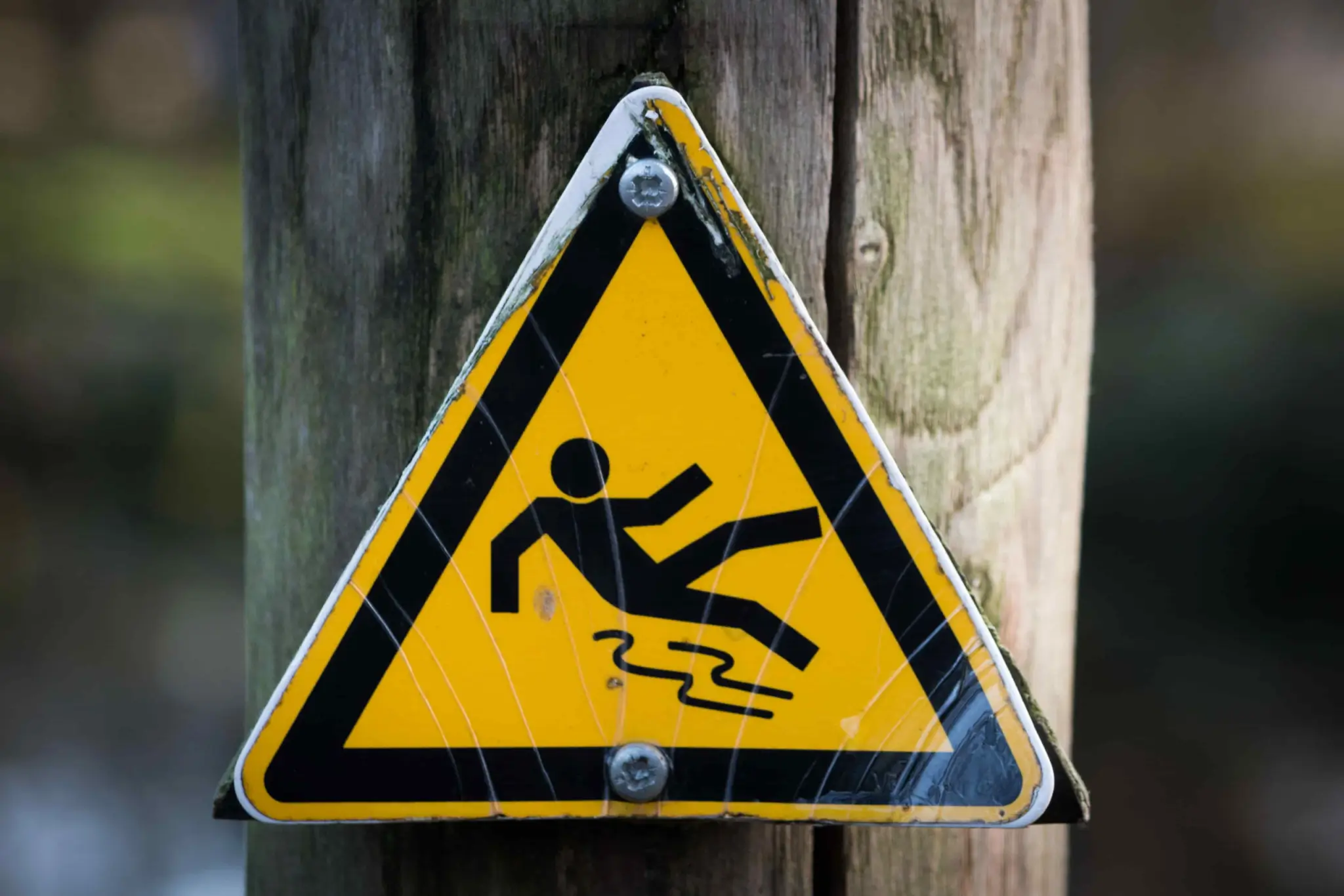 Yellow triangular warning sign attached to a wooden post, showing a stick figure slipping, indicating a slippery surface hazard. - Ghassemi Law Group Yellow triangular warning sign attached to a wooden post, showing a stick figure slipping, indicating a slippery surface hazard