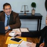 Three people in business attire have a meeting at a table with a laptop, documents, coffee cup, and scales of justice visible in the background. - Ghassemi Law Group Three people in business attire have a meeting at a table with a laptop, documents, coffee cup, and scales of justice visible in the background