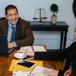 Three people sit at a meeting table with a laptop, documents, and coffee; one man smiles while others listen  A set of scales and a potted plant are on a desk in the background 