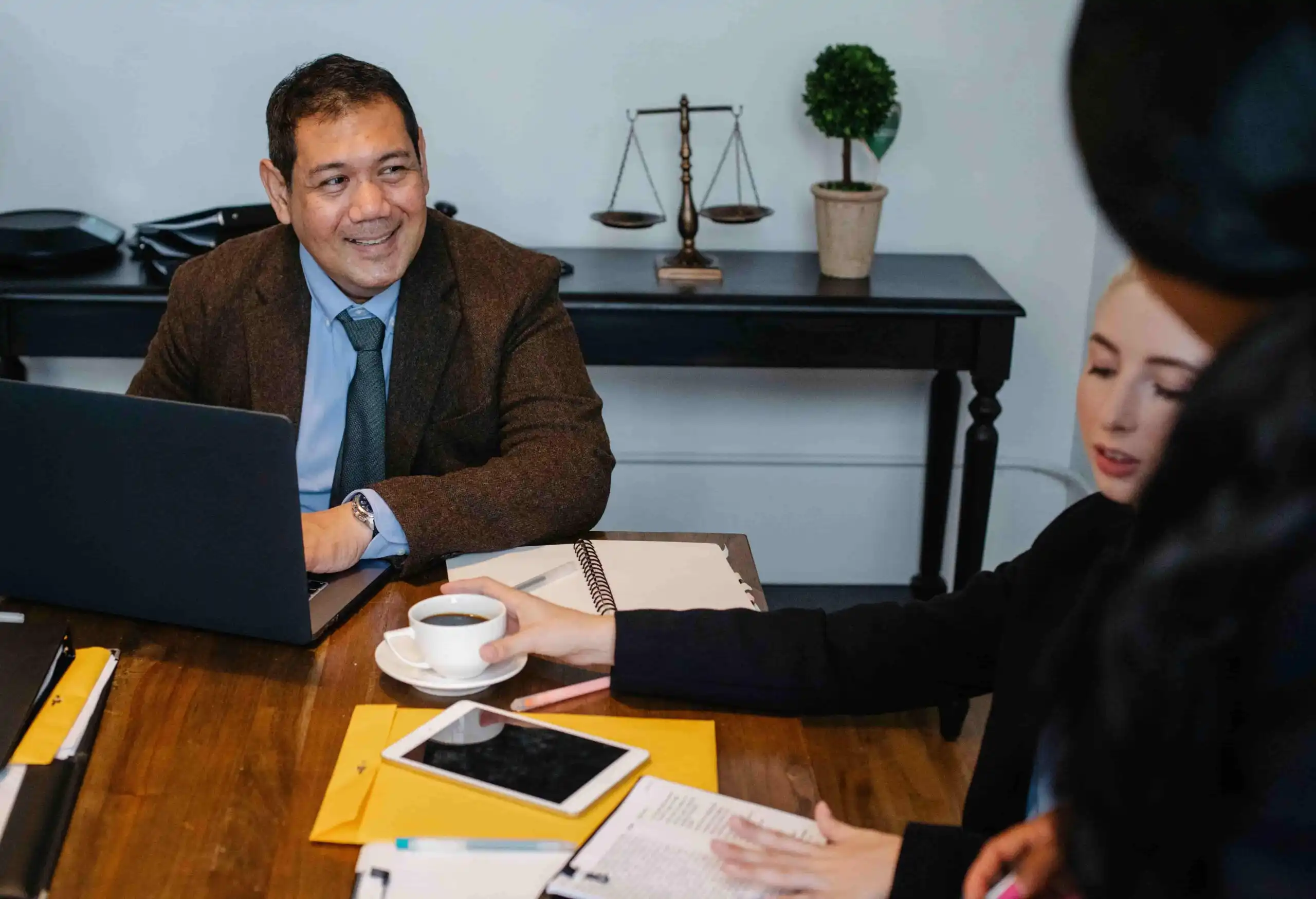 Three people sit at a meeting table with a laptop, documents, and coffee; one man smiles while others listen  A set of scales and a potted plant are on a desk in the background 