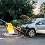 A silver car with a damaged front end is crashed into a bent streetlight on a road, with debris scattered around the scene 