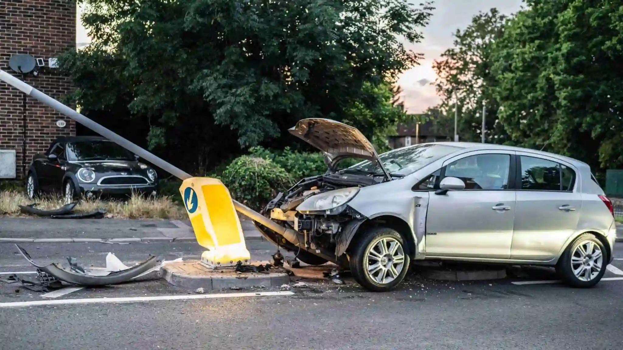 A silver car with a damaged front end is crashed into a bent streetlight on a road, with debris scattered around the scene 