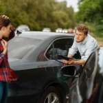 Two people stand next to damaged cars on a road, appearing to discuss a car accident  One person holds a clipboard, while the other touches their head  Trees are visible in the background 