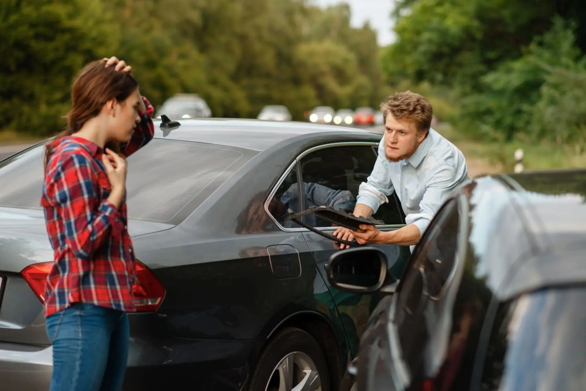 Two people stand next to damaged cars on a road, appearing to discuss a car accident. One person holds a clipboard, while the other touches their head. Trees are visible in the background. - Ghassemi Law Group Two people stand next to damaged cars on a road, appearing to discuss a car accident One person holds a clipboard, while the other touches their head Trees are visible in the background