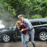 A man helps a woman away from a smoking, damaged SUV after a rollover accident  Both people appear unharmed but distressed  Trees and greenery are visible in the background 