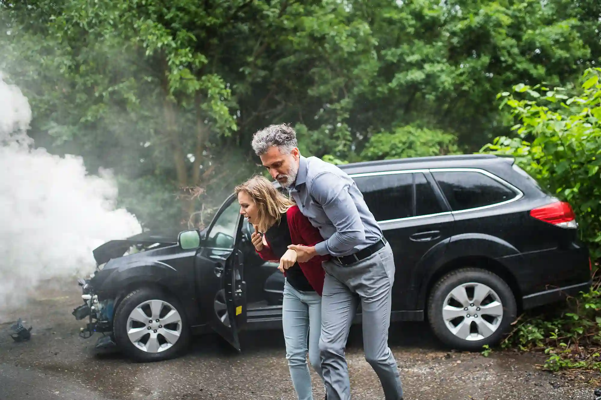 A man helps a woman away from a smoking, damaged SUV after a rollover accident. Both people appear unharmed but distressed. Trees and greenery are visible in the background. - Ghassemi Law Group - Ghassemi Law Group A man helps a woman away from a smoking, damaged SUV after a rollover accident Both people appear unharmed but distressed Trees and greenery are visible in the background