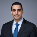 Man wearing a black suit, white shirt, and blue tie, facing the camera and smiling slightly, with a plain gray background—a professional look ideal for representing Ghassemi Law 