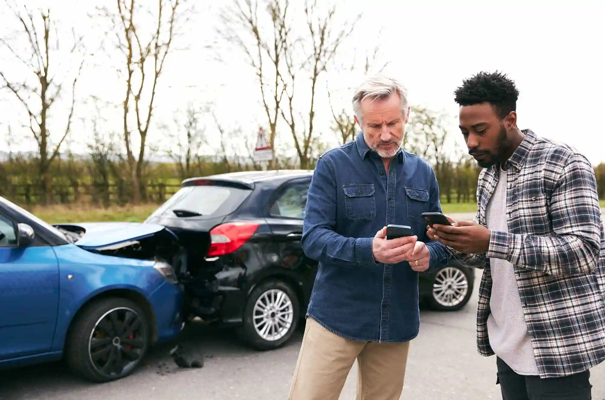 Two men stand next to damaged cars after an automobile accident, looking at their phones—possibly exchanging information or contacting assistance