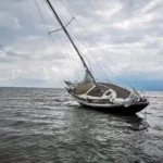 A sailboat is tilted to one side in choppy water under a cloudy sky, appearing damaged or abandoned 