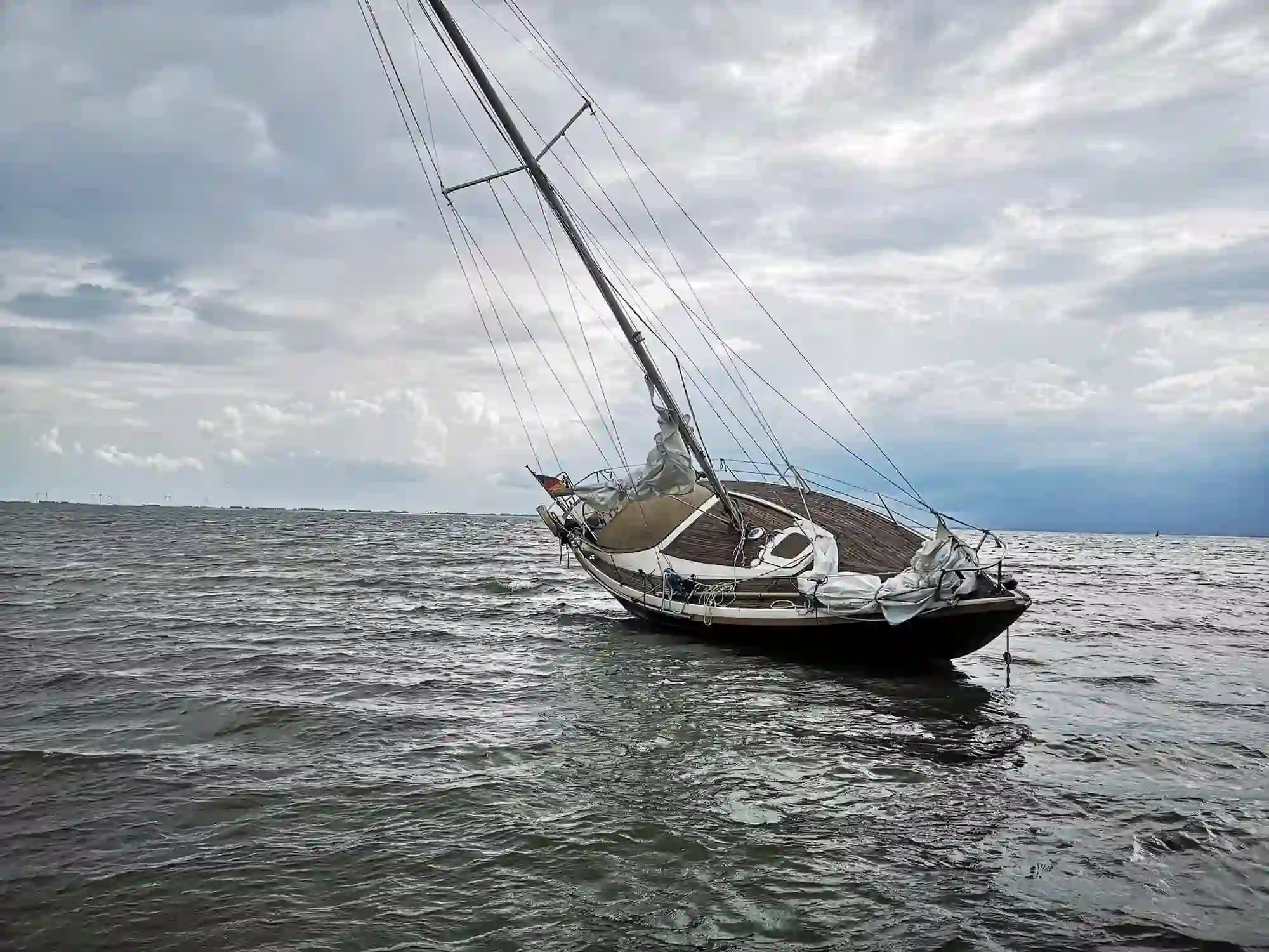 A sailboat is tilted to one side in choppy water under a cloudy sky, appearing damaged or abandoned 