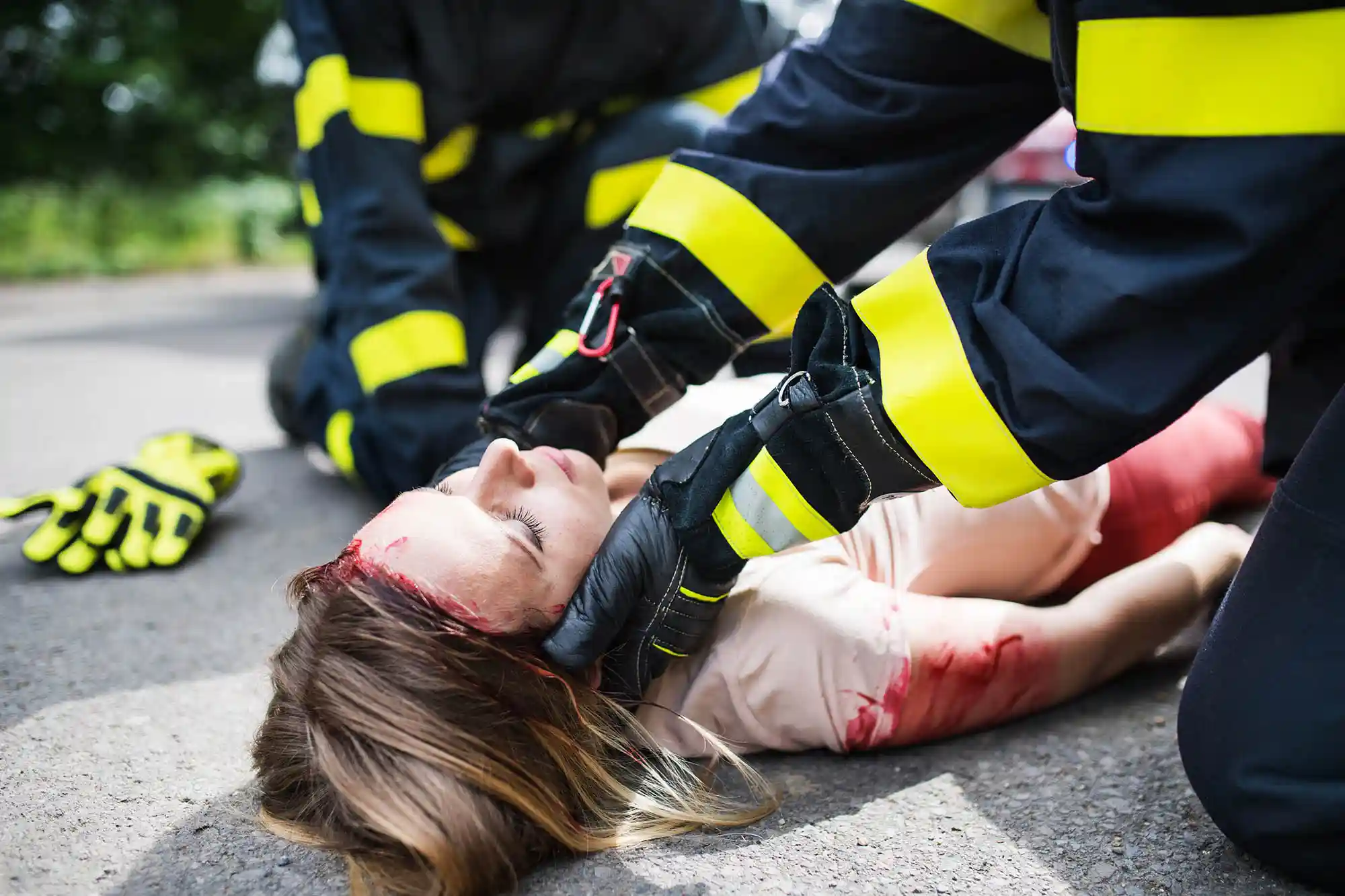 Two emergency responders tend to an injured woman lying on the ground with a head wound and blood on her arm, providing urgent care for what appear to be catastrophic injuries
