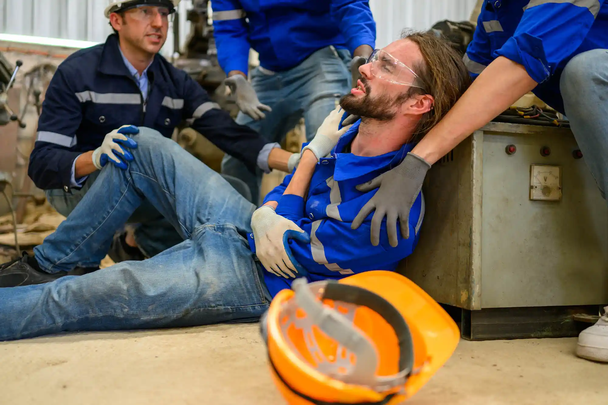 Three workers assist a colleague sitting on the floor, appearing injured—possibly due to construction injuries. A safety helmet lies nearby. Everyone is wearing gloves, glasses, and blue uniforms in an industrial setting. - Ghassemi Law Group - Ghassemi Law Group Three workers assist a colleague sitting on the floor, appearing injured—possibly due to construction injuries A safety helmet lies nearby Everyone is wearing gloves, glasses, and blue uniforms in an industrial setting