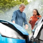 Two people stand between two cars with damaged front ends, appearing to discuss the accident  Smoke is visible, and trees are in the background, suggesting this may have been one of many distracted driver accidents 