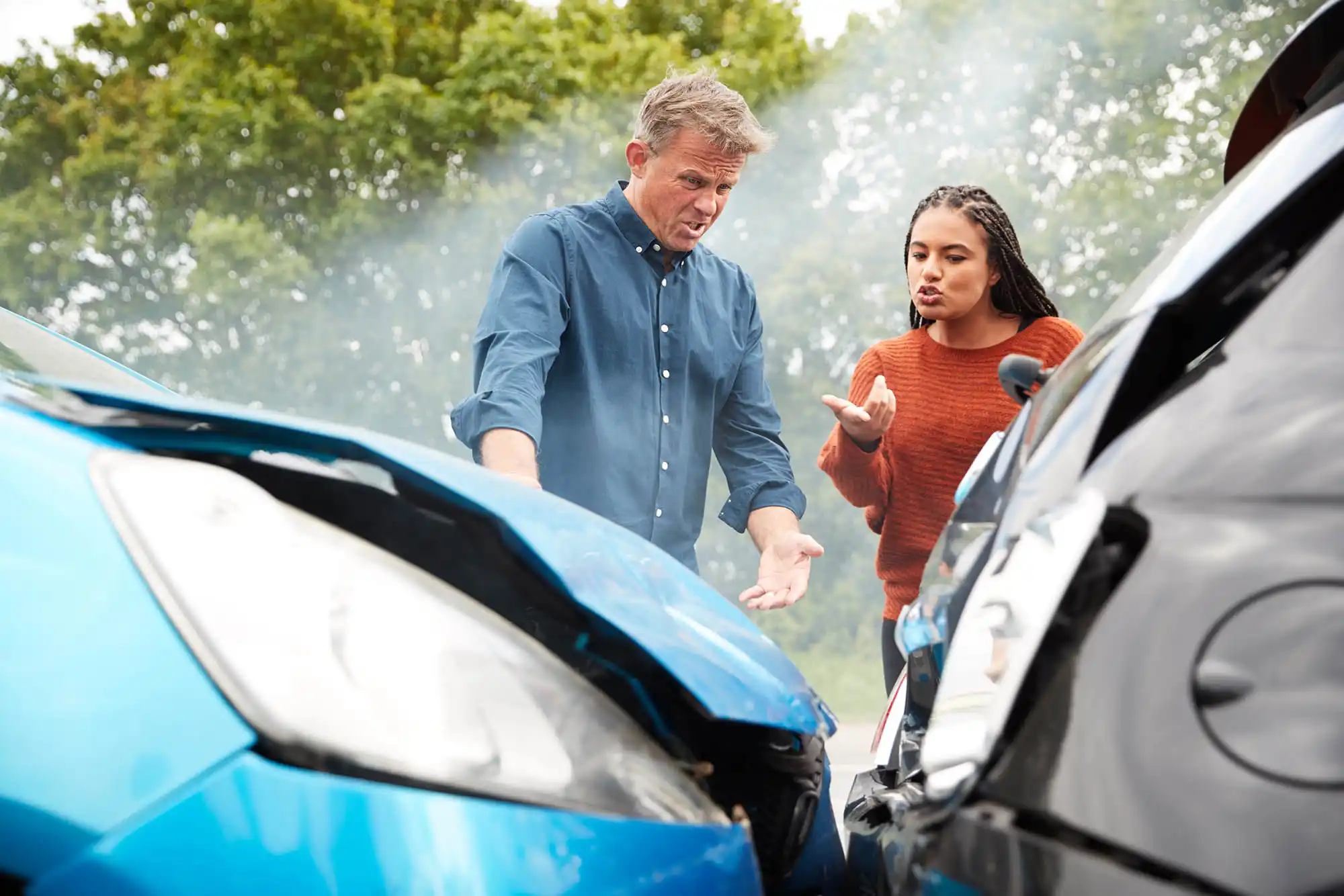 Two people stand between two cars with damaged front ends, appearing to discuss the accident  Smoke is visible, and trees are in the background, suggesting this may have been one of many distracted driver accidents 