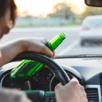 Person holding a green bottle while driving a car, with hands on the steering wheel and another vehicle visible through the windshield—highlighting the dangers of a potential drunk driving accident 