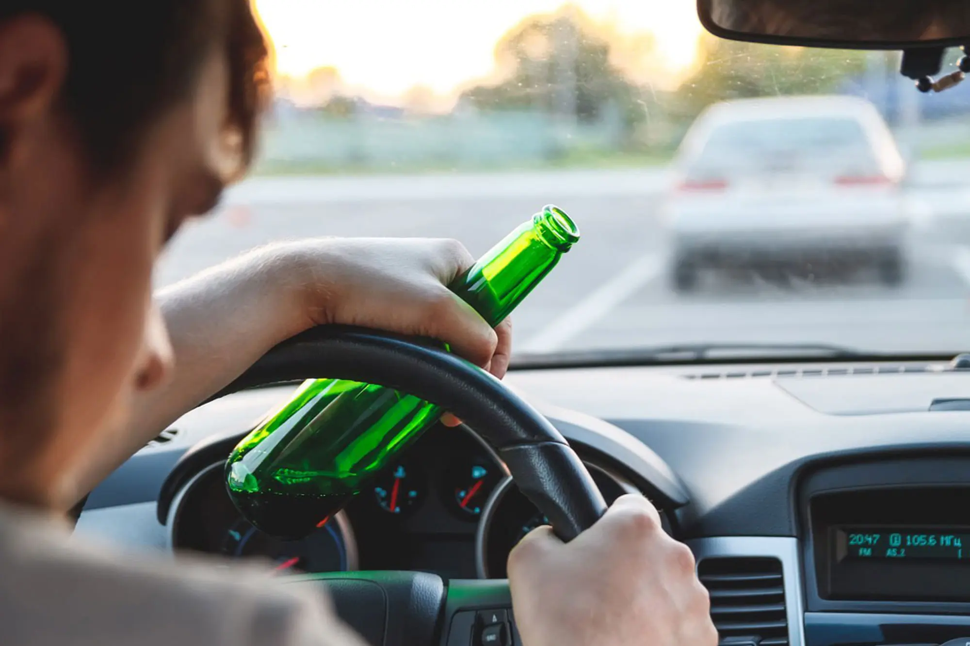 Person holding a green bottle while driving a car, with hands on the steering wheel and another vehicle visible through the windshield—highlighting the dangers of a potential drunk driving accident. - Ghassemi Law Group Person holding a green bottle while driving a car, with hands on the steering wheel and another vehicle visible through the windshield—highlighting the dangers of a potential drunk driving accident