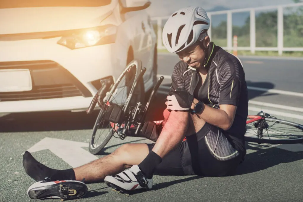 A cyclist wearing a helmet sits on the road holding his injured knee next to a fallen e-bike after an E-Bike Accident, with a white car nearby. - Ghassemi Law Group A cyclist wearing a helmet sits on the road holding his injured knee next to a fallen bicycle, with a car stopped behind him.