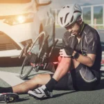 A cyclist sits on the road holding his injured knee next to a fallen bicycle after an E Bike accident, with a car stopped behind him 