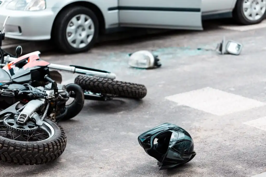 A motorcycle and a helmet lie on the road next to a car with an open door, with car parts scattered nearby.