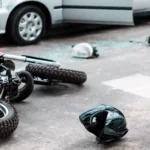 A motorcycle lies on its side near a helmet and broken car parts; a car with an open door is parked nearby 