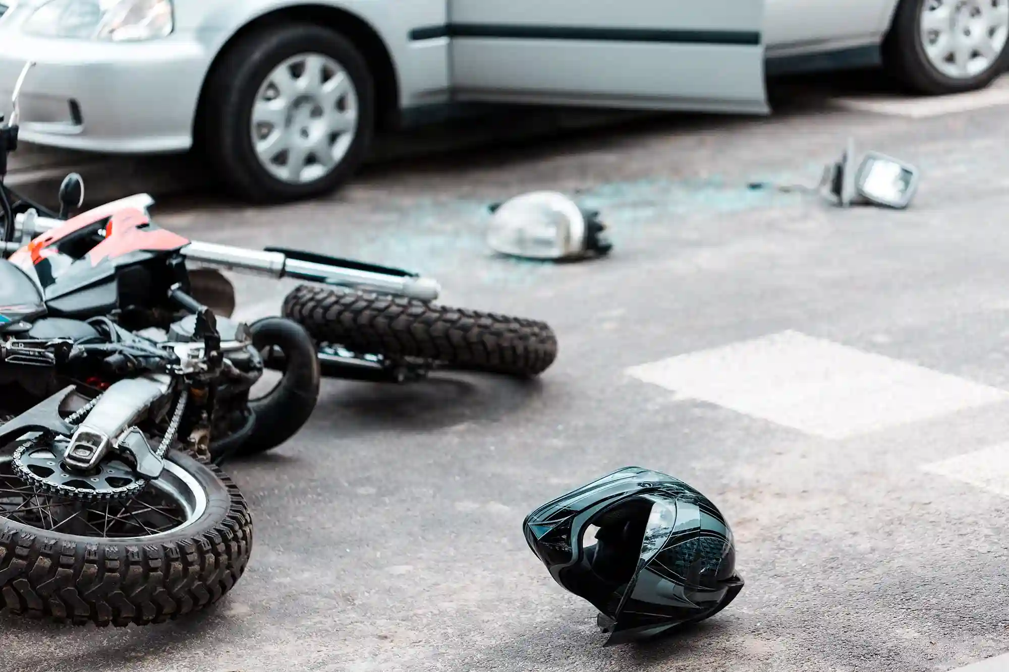 A motorcycle lies on its side near a helmet and broken car parts; a car with an open door is parked nearby