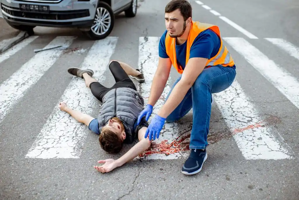 A man in a safety vest tends to an injured person lying on a crosswalk with visible blood, possibly after an accident.