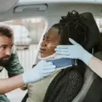 Two paramedics fit a neck brace on a woman seated in a car  The woman appears to be in pain, and both paramedics wear green uniforms and blue gloves 