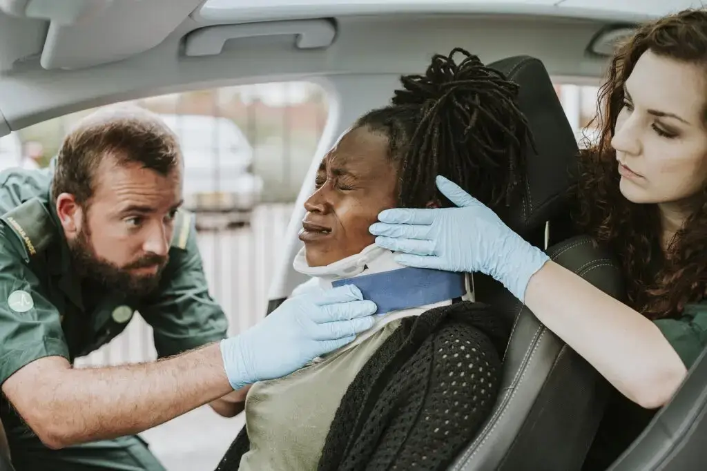 Two paramedics fit a neck brace on a woman seated in a car  The woman appears to be in pain, and both paramedics wear green uniforms and blue gloves
