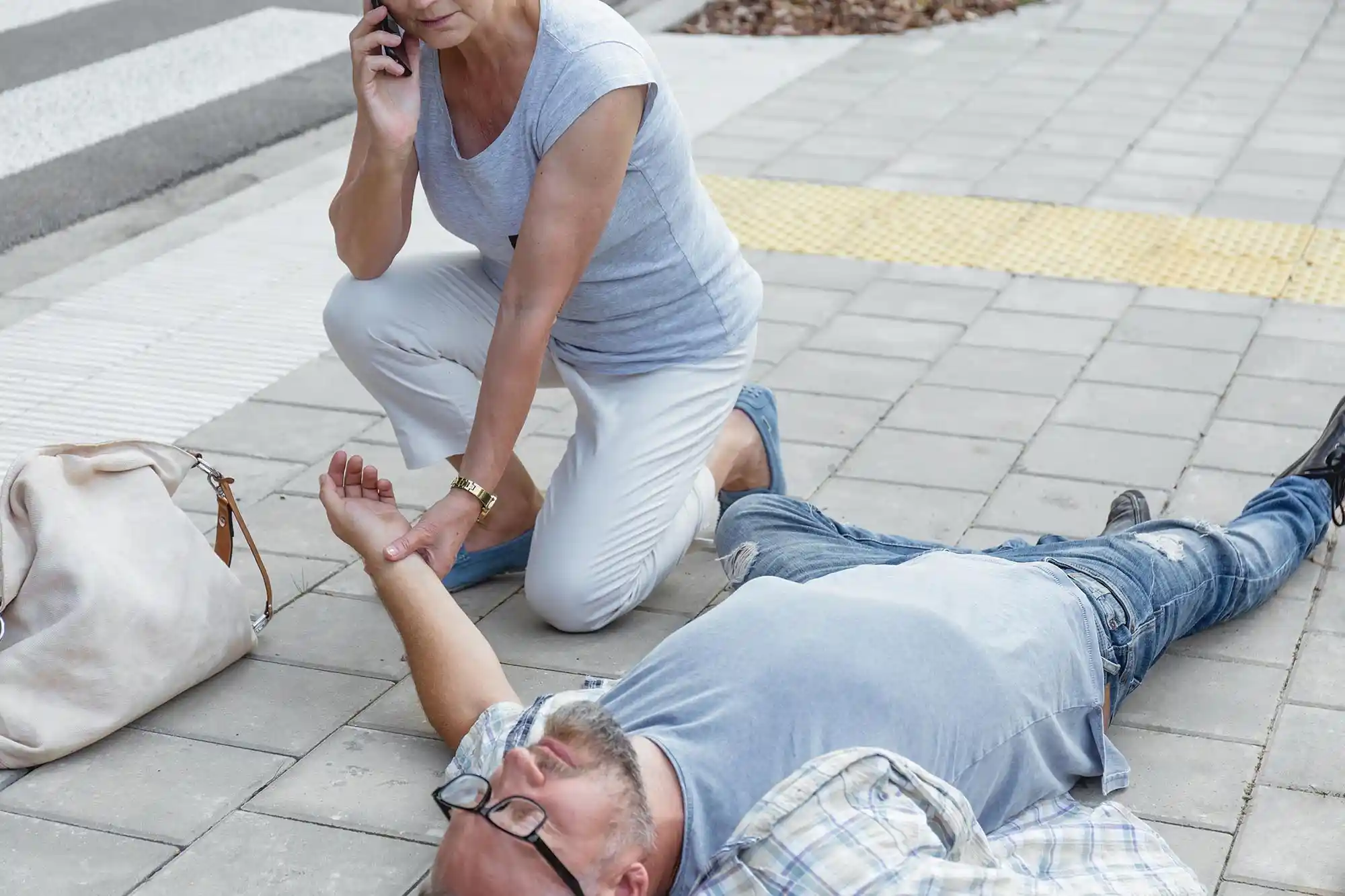 A woman kneels on a sidewalk outside a business, checking the pulse of a man lying unconscious while speaking on her phone—an urgent moment that may involve issues of premises liability