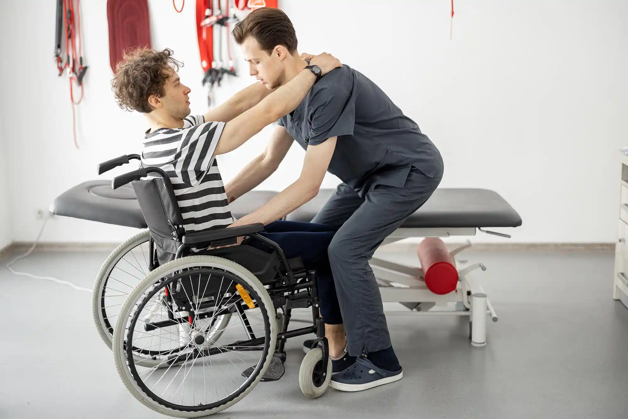 A physical therapist assists a man in a wheelchair with transfer exercises in a therapy room equipped with medical equipment, providing specialized care for individuals recovering from spinal cord injuries