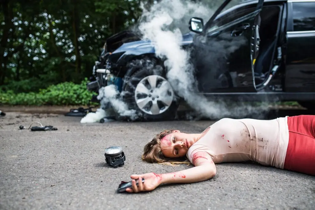 Injured woman lies on the road in front of a crashed, smoking vehicle with debris scattered around.