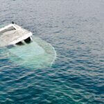 A partially submerged white boat is sinking in calm blue water, with only part of the cabin and hull visible above the surface 