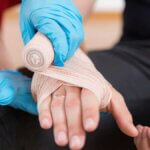 A person with a bandaged hand is receiving first aid from someone wearing blue gloves who is wrapping gauze around the injury. - Ghassemi Law Group A person with a bandaged hand is receiving first aid from someone wearing blue gloves who is wrapping gauze around the injury