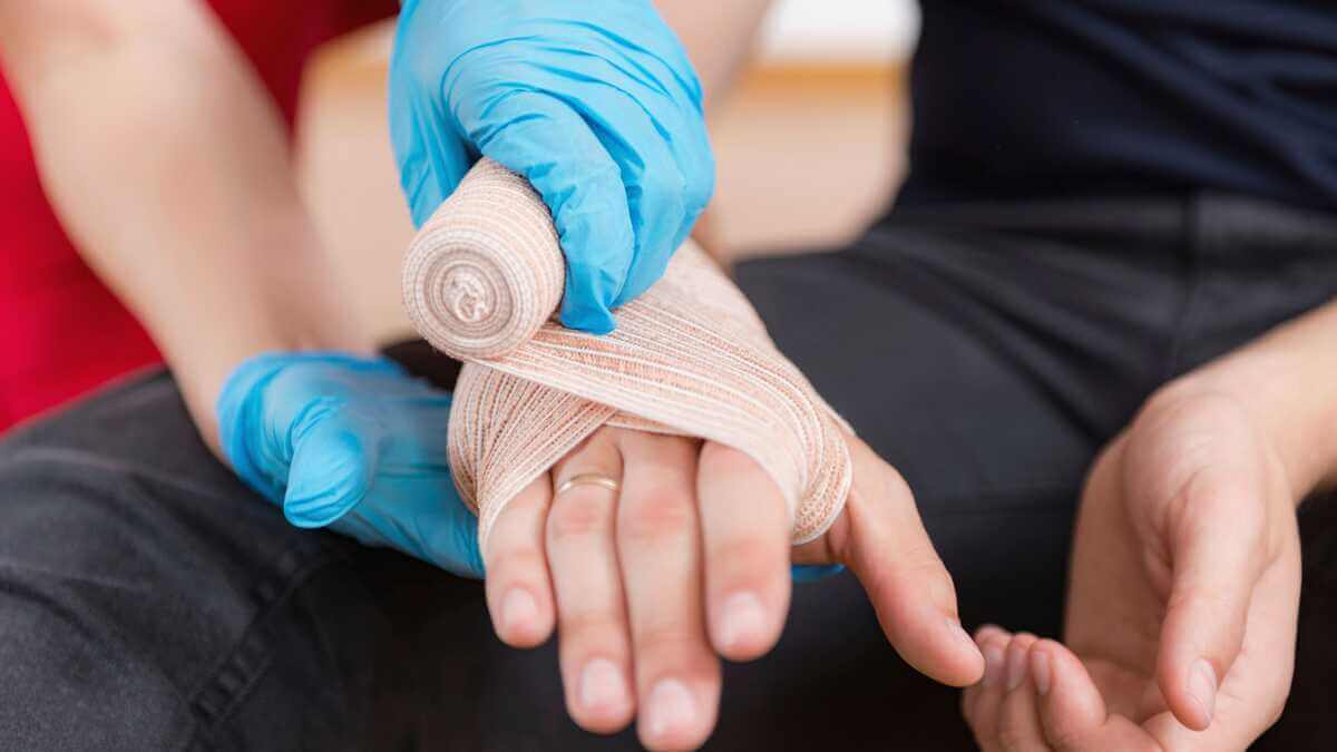 A person with a bandaged hand is receiving first aid from someone wearing blue gloves who is wrapping gauze around the injury. - Ghassemi Law Group A person with a bandaged hand is receiving first aid from someone wearing blue gloves who is wrapping gauze around the injury