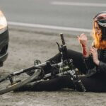 A person wearing a helmet sits on the ground next to a fallen bicycle, inspecting a bleeding arm, with a car parked nearby. - Ghassemi Law Group A person wearing a helmet sits on the ground next to a fallen bicycle, inspecting a bleeding arm, with a car parked nearby