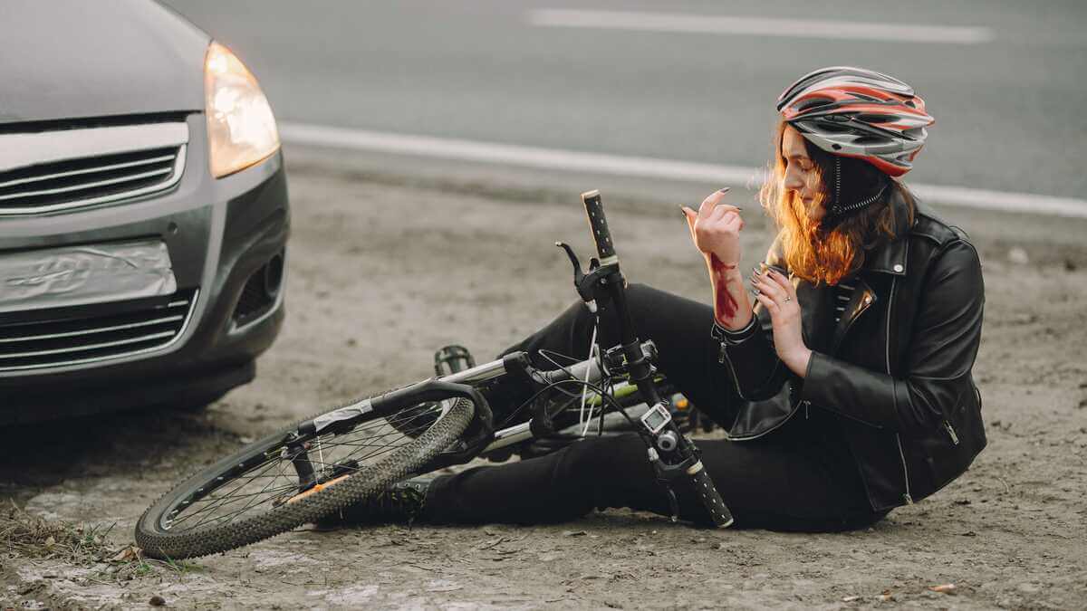 A person wearing a helmet sits on the ground next to a fallen bicycle, inspecting a bleeding arm, with a car parked nearby. - Ghassemi Law Group A person wearing a helmet sits on the ground next to a fallen bicycle, inspecting a bleeding arm, with a car parked nearby