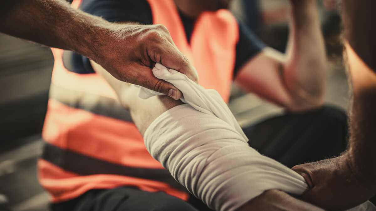 A person in a safety vest wraps a cloth bandage around another person’s injured forearm 