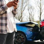 A person using a smartphone stands near two cars involved in a rear end collision on a road 