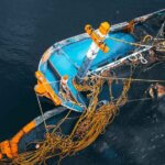 A partially sunken blue and orange boat with ropes and fishing nets floats in dark water, viewed from above 