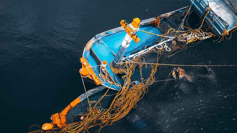 A partially sunken blue and orange boat with ropes and fishing nets floats in dark water, viewed from above. - Ghassemi Law Group A partially sunken blue and orange boat with ropes and fishing nets floats in dark water, viewed from above