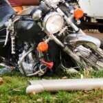 A motorcycle is lying on its side on grass near a downed metal pole, close to a white vehicle, suggesting a recent accident 