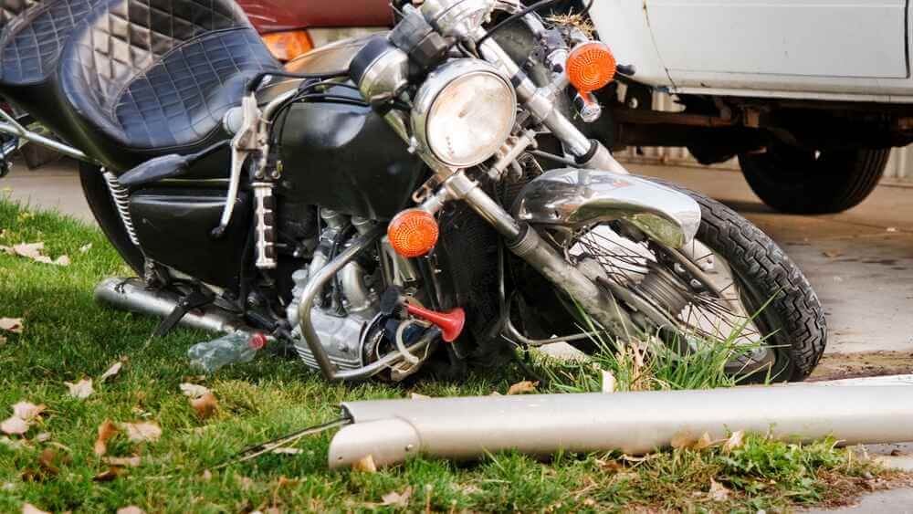 A motorcycle is lying on its side on grass near a downed metal pole, close to a white vehicle, suggesting a recent accident 