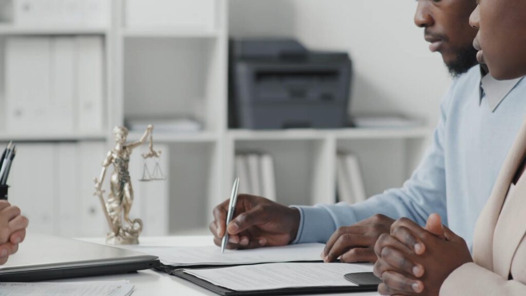 Two people sit at a desk reviewing and signing documents, with a Lady Justice statue and office shelves in the background.